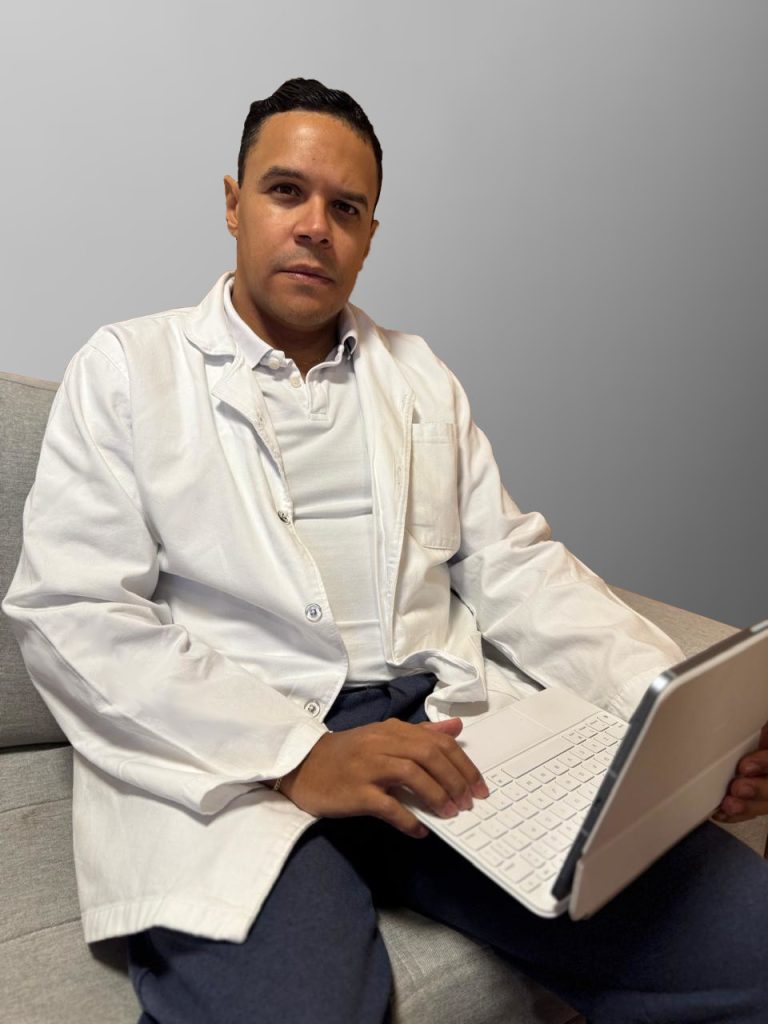Diego de jaleco branco sentado com notebook no seu colo olhando para a câmera.